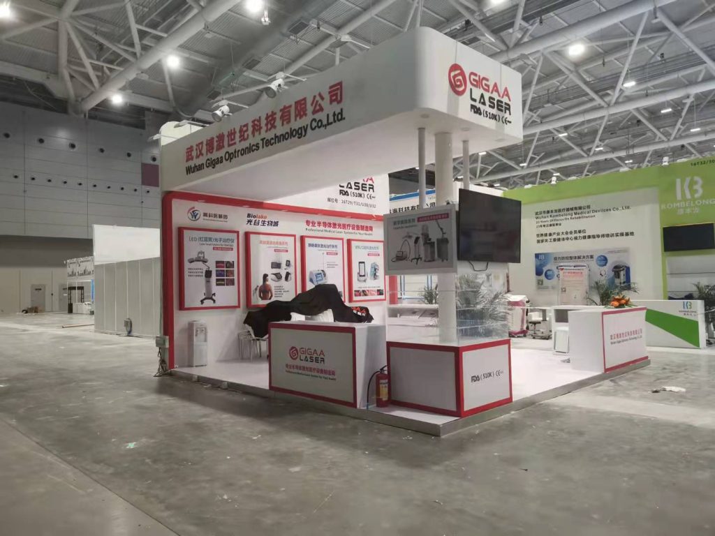 A wide shot of an uncompleted exhibition booth with white and red accents, tables covered, within a spacious convention hall.