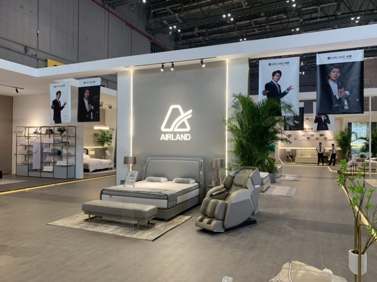 A sleek exhibition booth showcases a grey bed, a matching massage chair, and a glowing logo on the wall. Banners of a man in a suit hang above.
