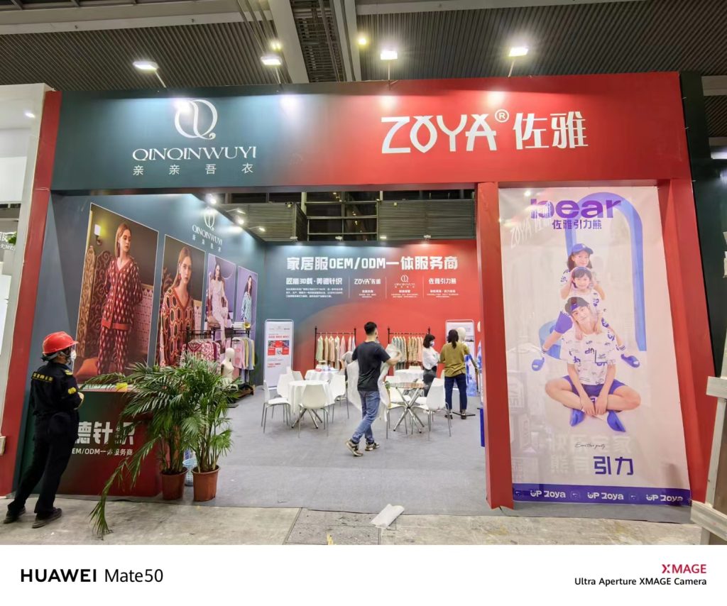 A large exhibition booth in red and green with clothing displays. People are walking inside, and models are shown on posters featuring adult and children's apparel.