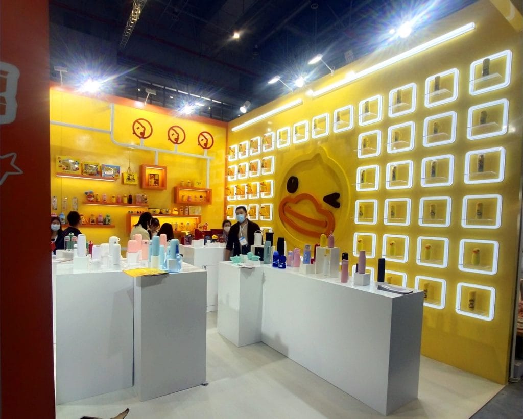 Inside the yellow booth, white pedestals display colorful bottles. People view products near a large duck logo.