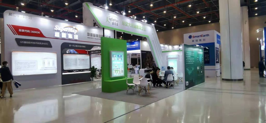 Real-life trade show booth, white and green, with an angled arch. People are seated inside, surrounded by informational displays, within a busy exhibition hall.