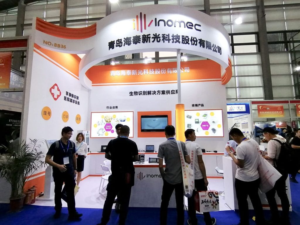 Attendees interact at an orange and white exhibition booth showcasing products on multiple screens within a bustling trade show hall.