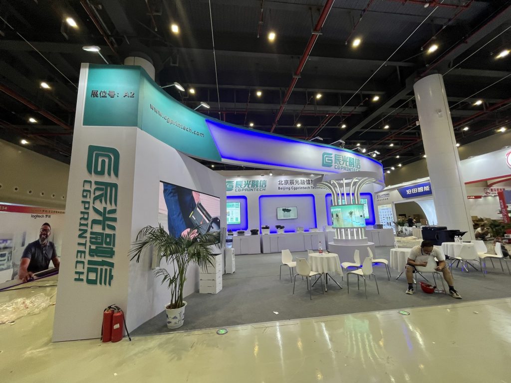 A brightly lit exhibition booth features a curved archway, display screens, and a central area with tables.