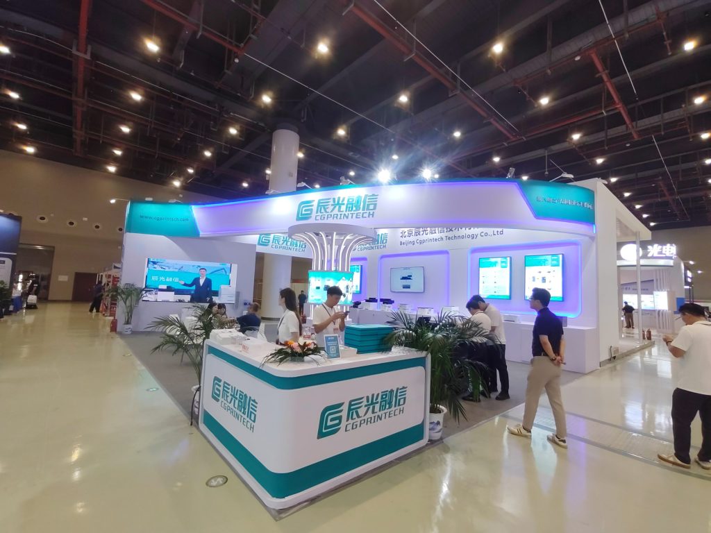 A busy exhibition booth with many people interacting at counters, tables, and product displays under a large, curved sign.