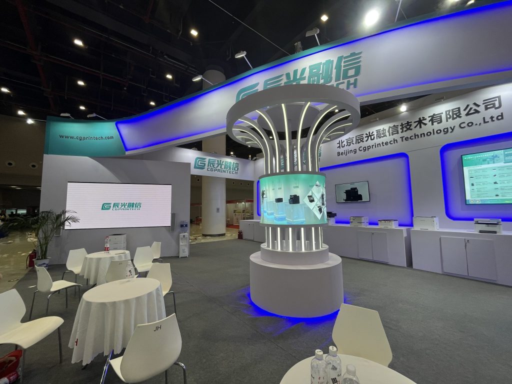 An exhibition booth with a striking cylindrical glowing display, surrounded by multiple white tables and chairs, showing products on wall screens.