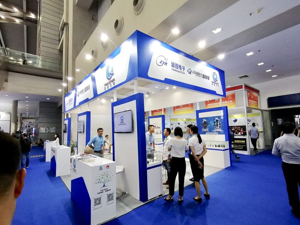 People gather at a blue and white exhibition booth, interacting and viewing displays of electronic components. A man in a blue shirt engages with attendees.