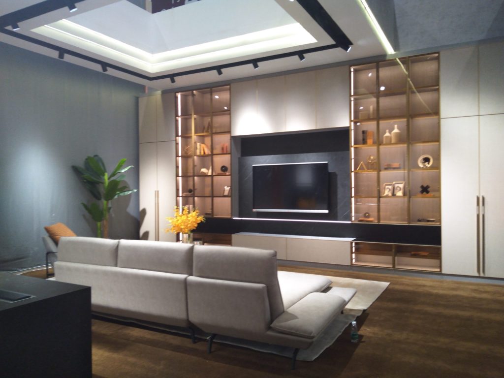 A contemporary living room with a light grey sectional sofa, a wall-mounted TV, and illuminated display shelves filled with decor.