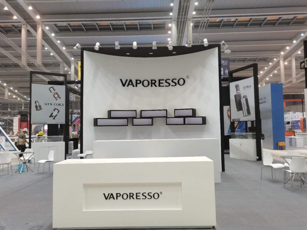 A white trade show booth with a large central sign, product display shelves, and a reception desk in a spacious convention hall.