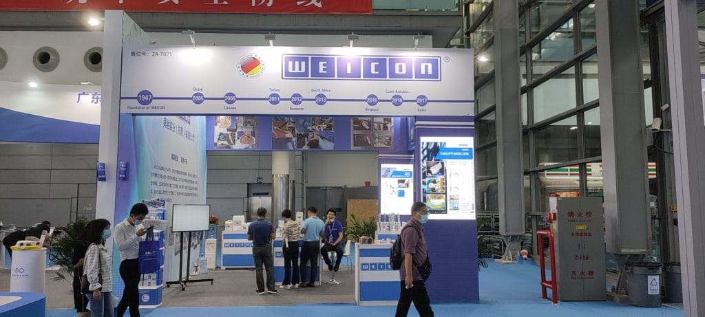 Visitors gather at a white and blue exhibition booth featuring product displays in a convention center.