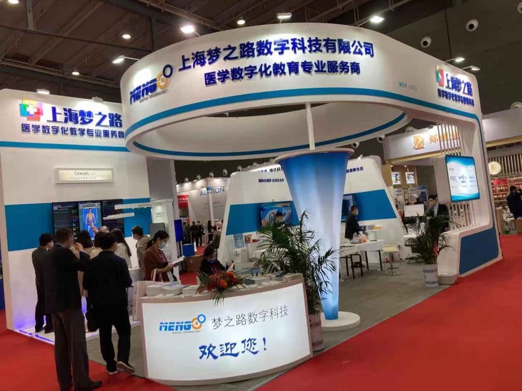 Many attendees gather around a large white and blue exhibition booth with a central reception desk at a trade show.