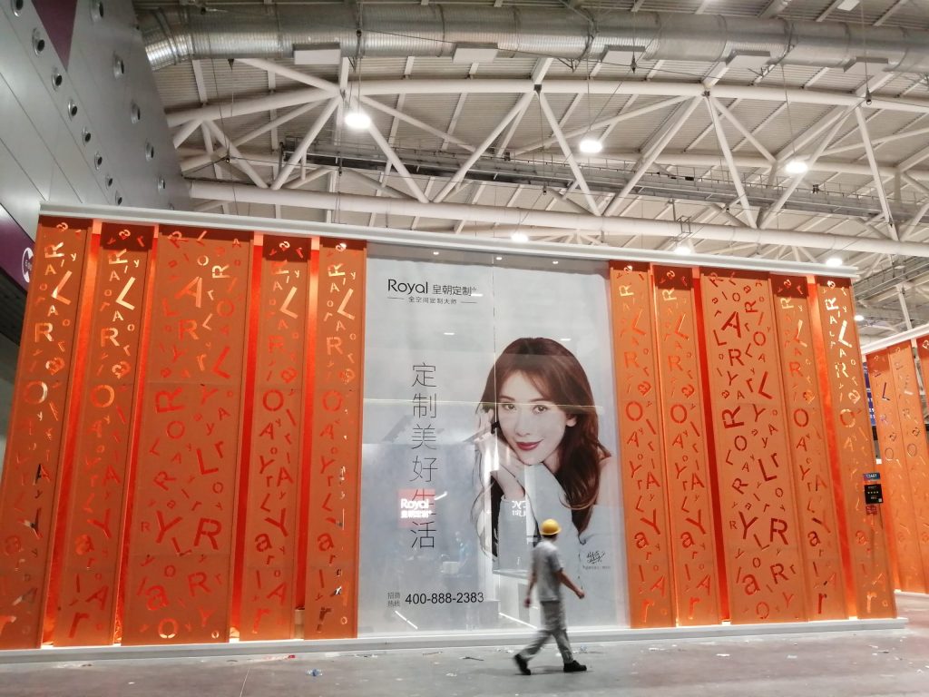 A worker in a yellow hard hat walks past an exhibition display wall with orange panels and a large portrait of a woman.