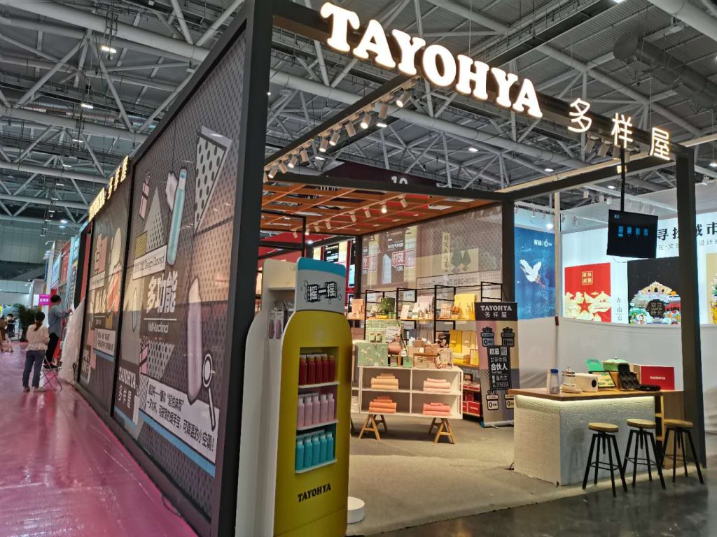 Side view of an exhibition booth with graphic walls and a yellow product display stand. Inside, shelves are filled with goods, and visitors are present.