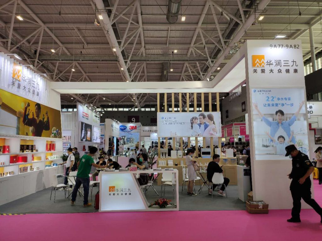 A bustling trade show floor with many visitors gathered around a brightly lit exhibition booth. A security guard walks in the foreground.