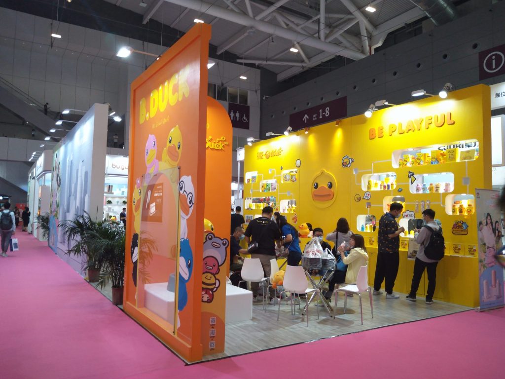A busy trade show booth with an orange arch entrance and a yellow wall displaying products, surrounded by many attendees.