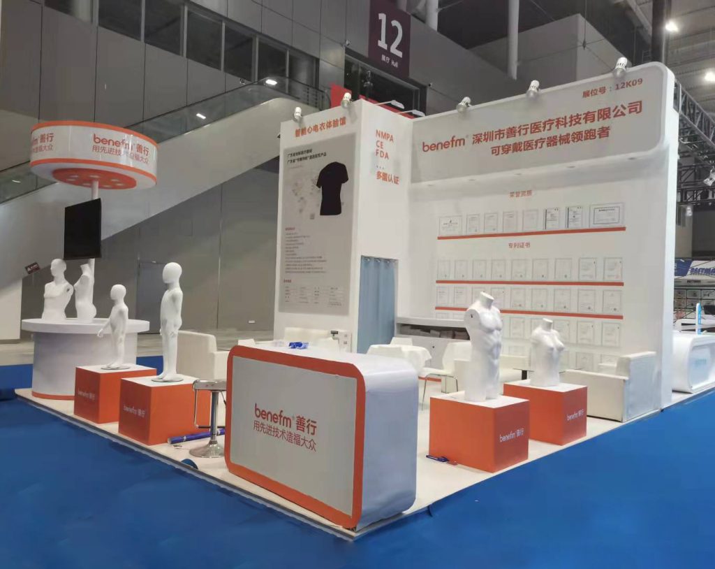 White and orange trade show booth on blue carpet, with diverse mannequins, product details, and framed certificates on walls.