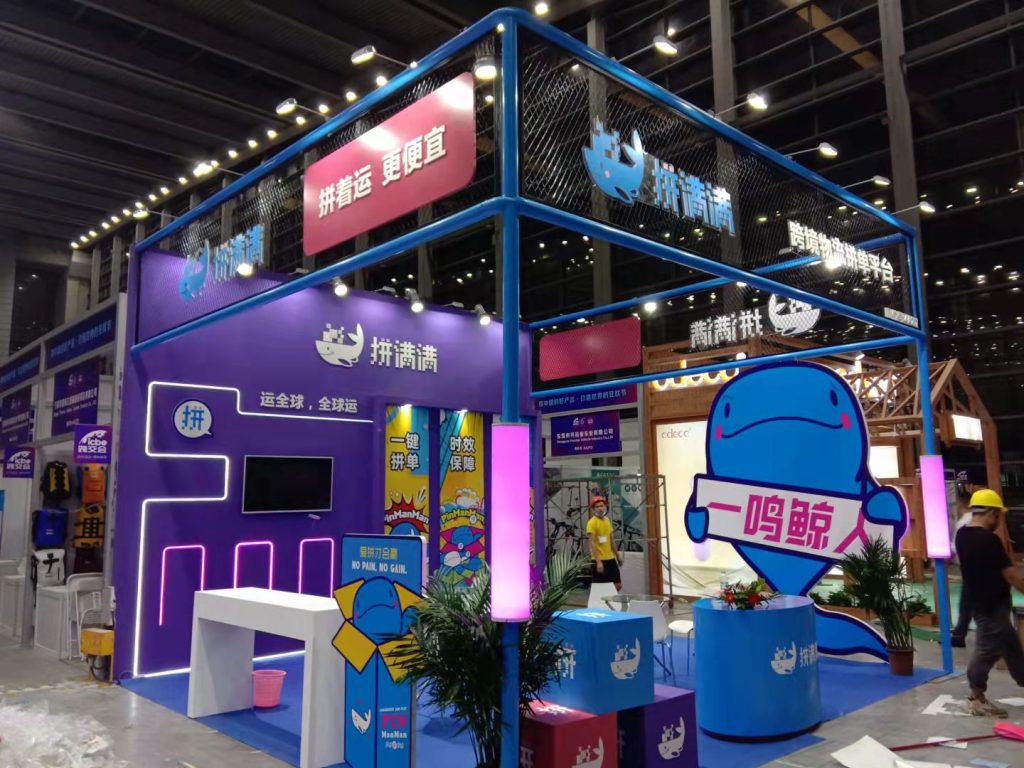 Exhibition booth featuring a prominent blue whale mascot cutout, counters, branded boxes, neon lighting, and staff in an active event setting.