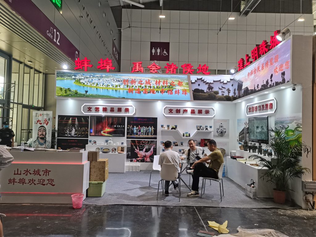 A bustling trade show booth displays diverse cultural content on multiple screens and products. People are conversing at a table inside the booth.