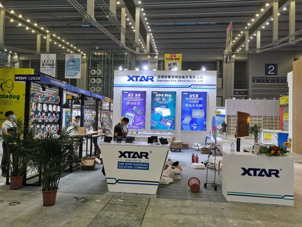 A partially constructed exhibition booth with blue display panels, white counters, and a person standing behind one. Boxes and equipment are on the concrete floor.
