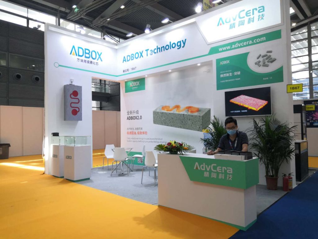 An operational trade show booth with a person in a mask behind the counter. Displays show ceramic components, alongside potted plants, chairs, and colorful flooring.