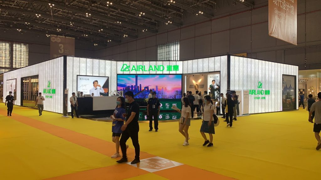 A prominent exhibition booth with glowing white walls and large display screens features various graphics. Many people stroll past the booth on a wide yellow carpet.
