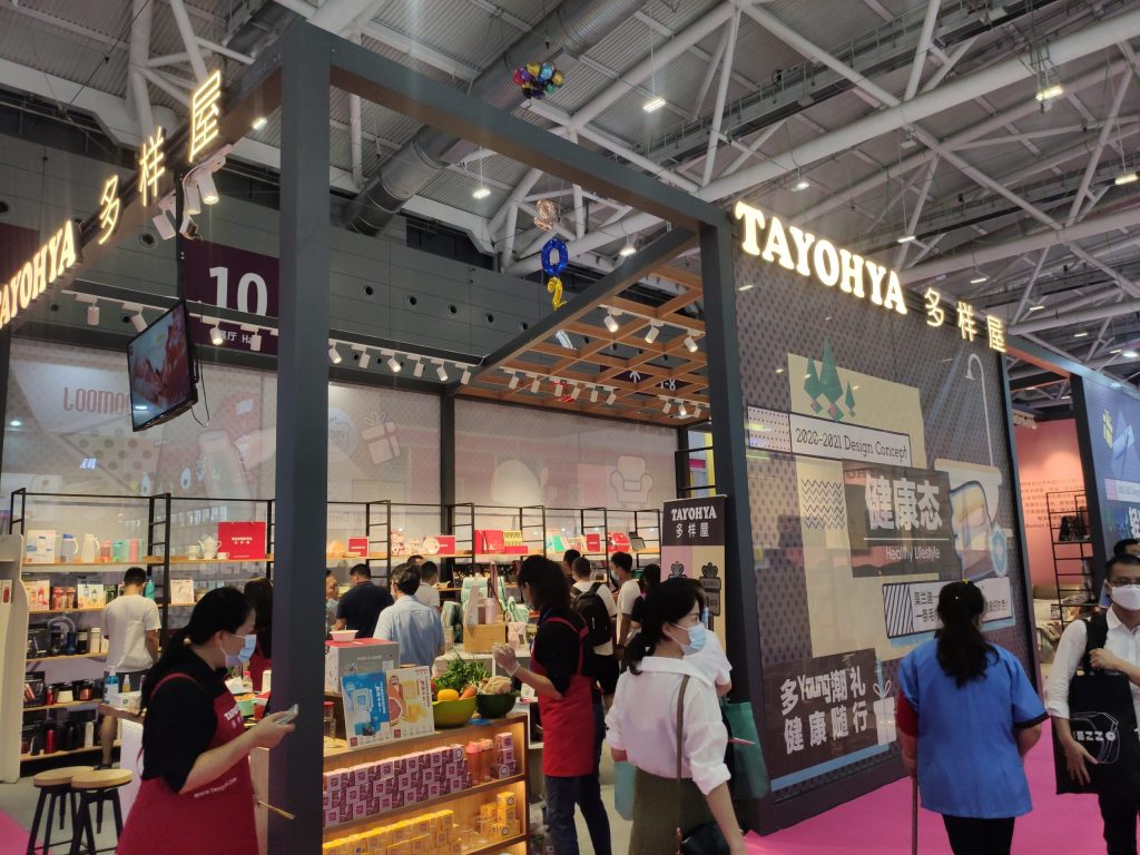 Wide view of a bustling exhibition booth with a pink floor. Many masked attendees and staff in red aprons move through the product displays.
