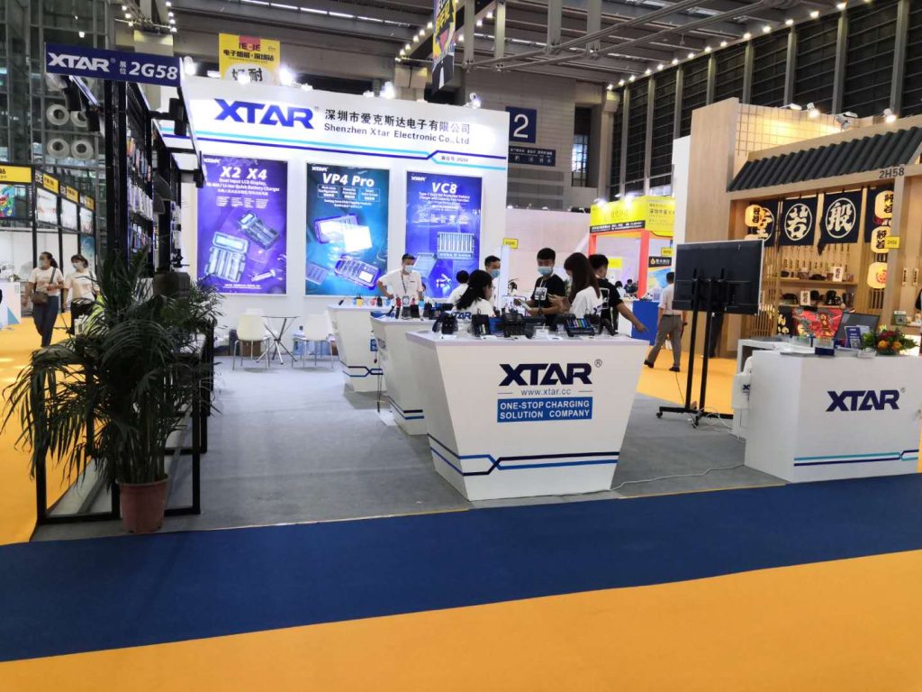 A bustling exhibition booth with blue display panels, white counters, and products on display. Multiple attendees and staff are present on the blue and yellow carpet.