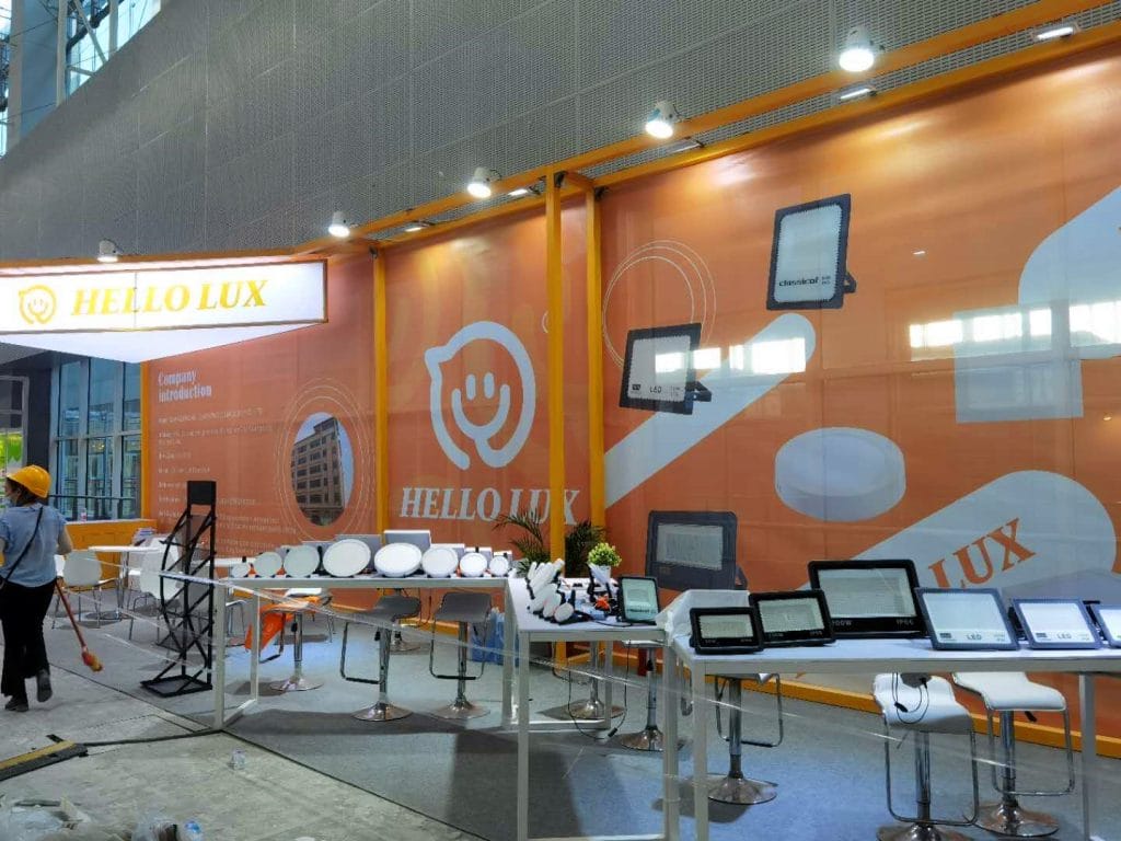 An orange and white exhibition booth displaying various lighting products on tables. A person in a hard hat walks past.
