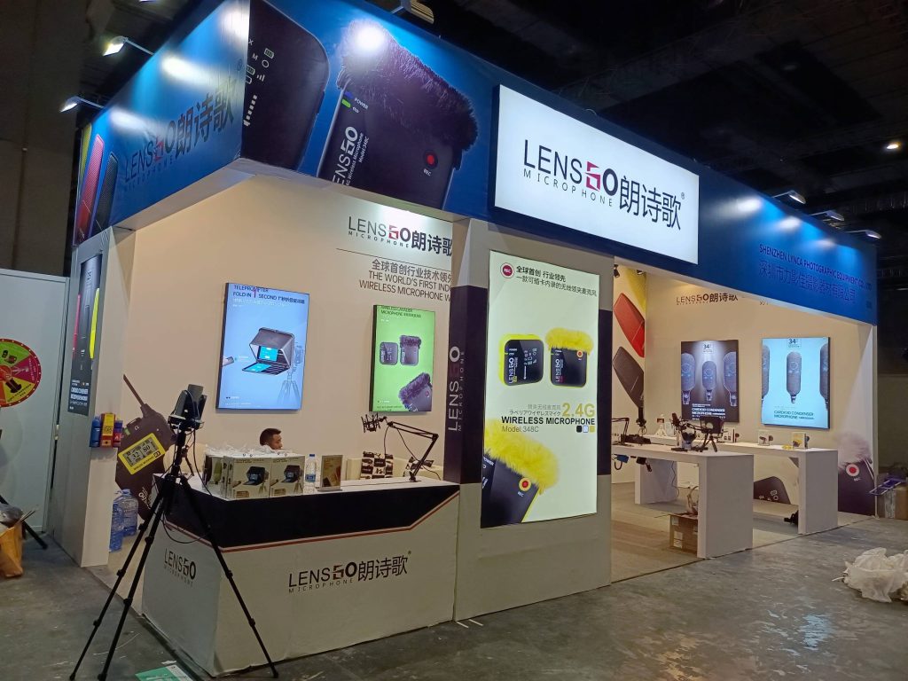 Angled view of a busy exhibition booth with masked visitors, a reception desk, and bright vertical displays showcasing various microphones.
