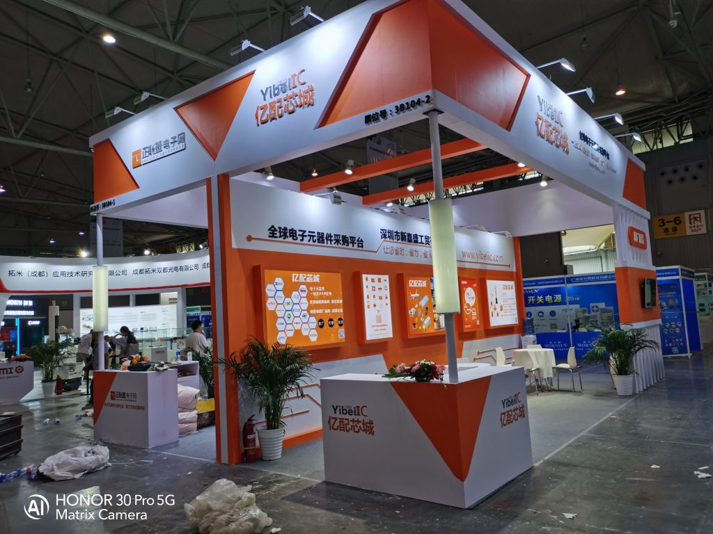 An orange and white exhibition booth with displays, seating, and decorative plants, set up in a large, busy trade show hall.