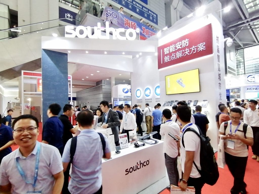 A busy trade show booth with many attendees interacting at white counters and viewing digital displays, under an arched sign in an exhibition hall.