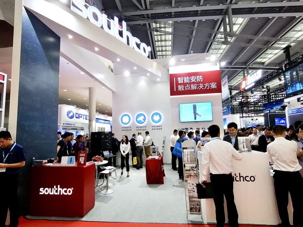 An active exhibition booth with attendees gathered around white and red counters, looking at product displays and screens within a large convention center.