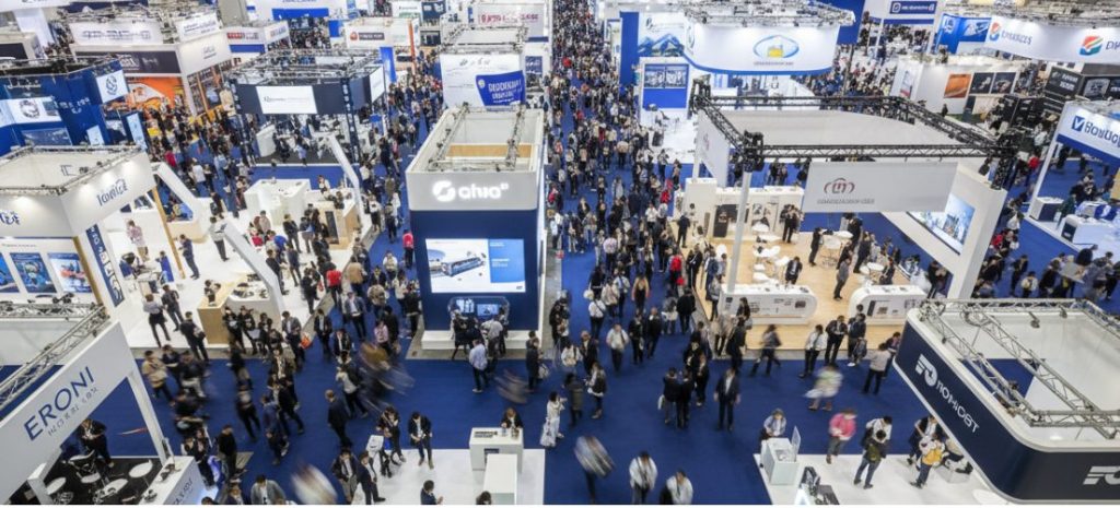 A bird's-eye view of a bustling international trade show, with dense crowds navigating blue-carpeted aisles between branded exhibition booths, showcasing a lively professional networking atmosphere.