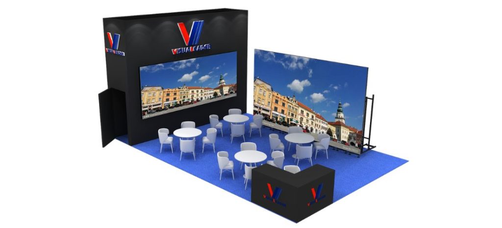 Event booth with large screens displaying a town square. Multiple white tables and chairs are arranged on a blue carpet, with a black counter in the foreground.