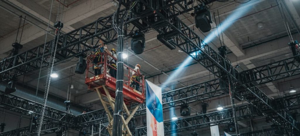 A busy exhibition construction site during setup day, workers installing booth structures, visible power cables on the floor, LED screens being assembled, slight tension in the environment, industrial lighting, realistic, documentary style, high detail, cinematic lighting, shallow depth of field